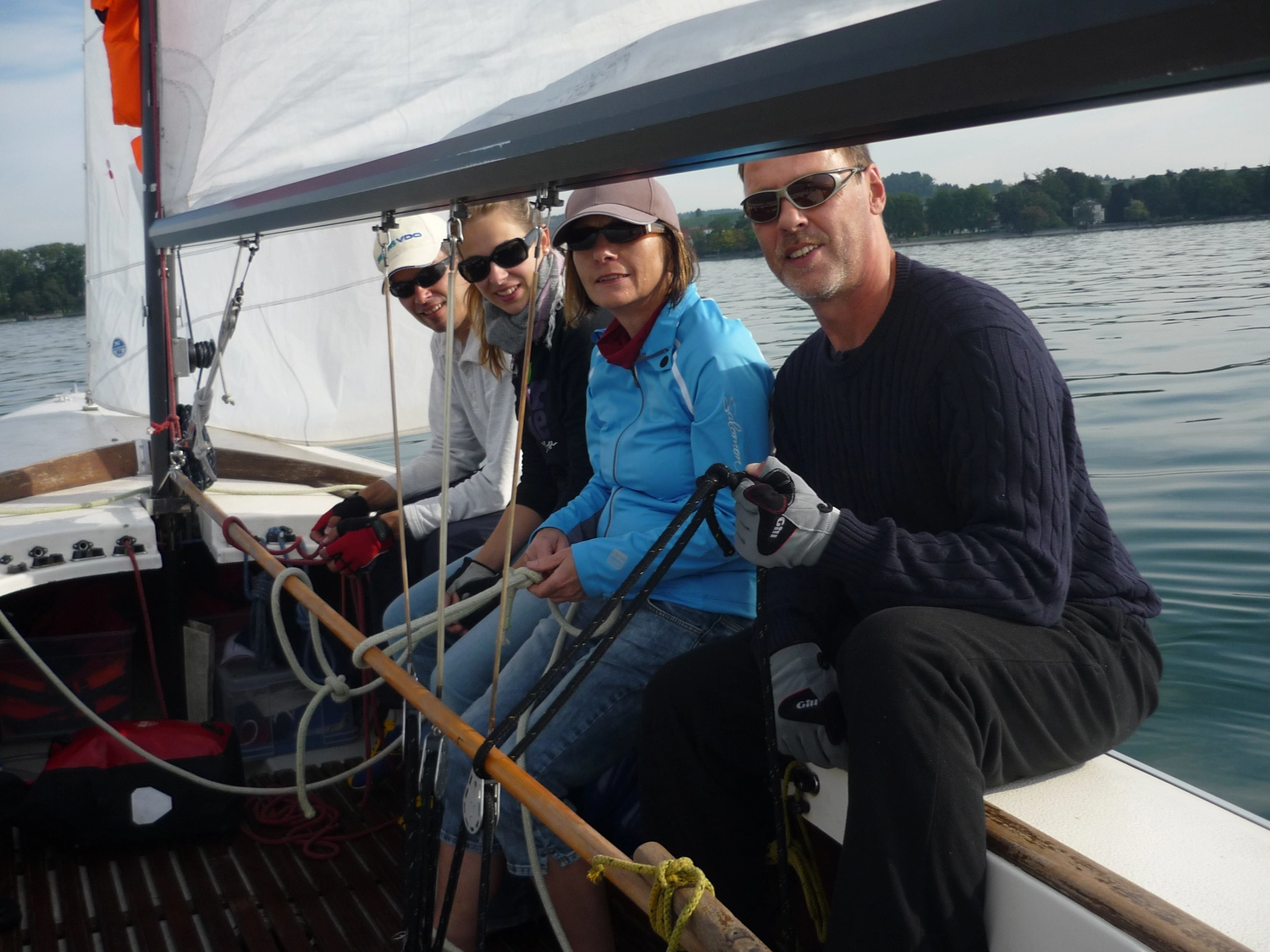 Motorboot-u. Segelschule Ravensburg - Wassersportausbildung und lizenzierung - Eine Gruppe von vier Personen lächelt, während sie einen Segeltag auf einem ruhigen Gewässer in einem Boot genießen.
